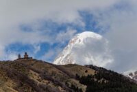 Kemegahan Gereja Gergeti Trinity yang berdiri kokoh di atas perbukitan hijau dengan latar belakang lanskap dramatis Pegunungan Kaukasus di kawasan Stepantsminda, Georgia.