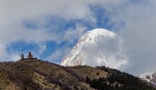 Kemegahan Gereja Gergeti Trinity yang berdiri kokoh di atas perbukitan hijau dengan latar belakang lanskap dramatis Pegunungan Kaukasus di kawasan Stepantsminda, Georgia.