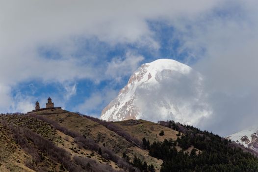 Kemegahan Gereja Gergeti Trinity yang berdiri kokoh di atas perbukitan hijau dengan latar belakang lanskap dramatis Pegunungan Kaukasus di kawasan Stepantsminda, Georgia.