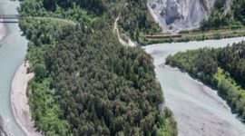 Sebuah kereta regional berwarna merah melintasi lembah hijau dengan latar tebing terjal dan puncak pegunungan Alpen Swiss yang megah.