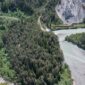 Sebuah kereta regional berwarna merah melintasi lembah hijau dengan latar tebing terjal dan puncak pegunungan Alpen Swiss yang megah.