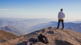 Seorang pelancong menikmati ketenangan pemandangan alam pegunungan yang sejuk dan asri, mencerminkan maraknya tren *coolcation* dan *slow travel* di kalangan wisatawan global.