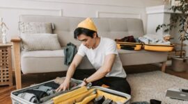 Seorang pelancong sedang menata pakaian secara rapi dengan teknik gulung ke dalam koper berukuran sedang untuk efisiensi ruang perjalanan.