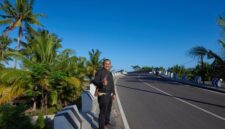 Sebuah kendaraan melintasi jalur pesisir berlatar pemandangan laut dan bukit hijau, menggambarkan pesona perjalanan darat yang menenangkan di Indonesia.