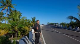 Sebuah kendaraan melintasi jalur pesisir berlatar pemandangan laut dan bukit hijau, menggambarkan pesona perjalanan darat yang menenangkan di Indonesia.